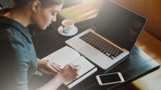 Candidate assise dans un café qui écrit le plan de sa lettre de motivation à la main devant son ordinateur