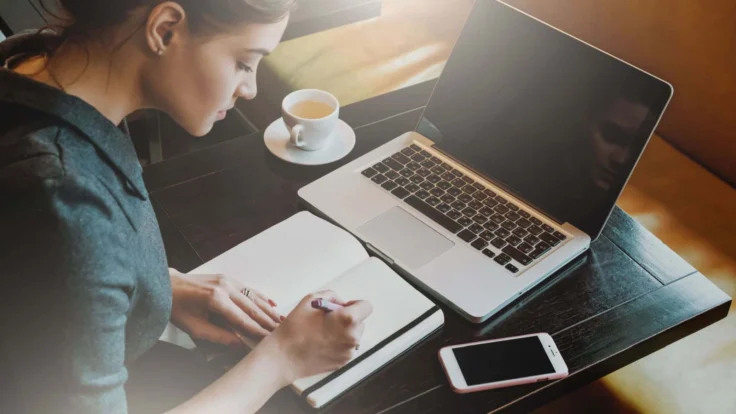 Candidate assise dans un café qui écrit le plan de sa lettre de motivation à la main devant son ordinateur